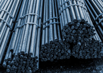 Bundles of steel rebar are stacked together, with the ends facing outward. The rods are tied with metal straps, displaying a textured, cylindrical surface. The image is captured with a blue tint, highlighting the industrial setting. | FintechZoom
