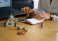 A person works at a wooden desk with a laptop, calculator, and notebook. A small house model and stacks of coins suggest budgeting for mortgage down payments. A pen and glass of water are also on the desk, completing the scene of financial planning. | FintechZoom