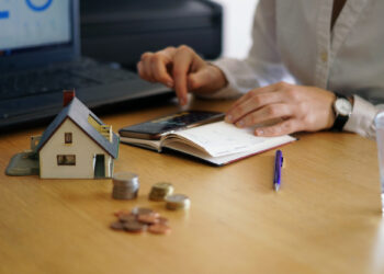 A person works at a wooden desk with a laptop, calculator, and notebook. A small house model and stacks of coins suggest budgeting for mortgage down payments. A pen and glass of water are also on the desk, completing the scene of financial planning. | FintechZoom
