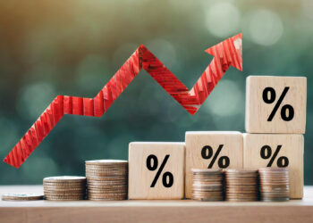 Stacks of coins and wooden blocks with percentage symbols show financial growth. A red zigzag arrow above the stacks points upward, indicating an increase. The background is blurred for focus on the coins and blocks. | FintechZoom