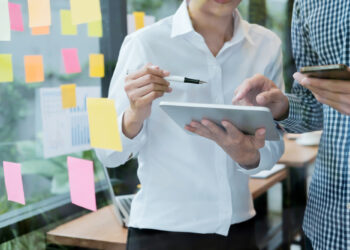 Two people manage multiple projects effectively in an office surrounded by colorful sticky notes on a glass wall. One points at a tablet with a pen, while the other holds a phone, seamlessly coordinating tasks. A laptop sits ready on the wooden table in the background. | FintechZoom