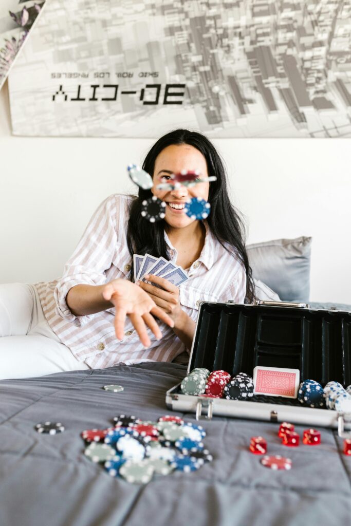 A person on a bed is joyfully tossing poker chips in the air. They hold playing cards in one hand and an open poker set with more chips and dice is nearby. A poster of a city skyline is on the wall in the background. | FintechZoom