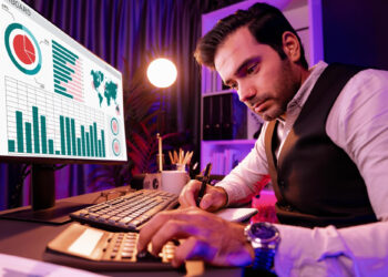 A man in a vest is focused on a computer screen displaying graphs and charts related to online casino games. He's typing on a calculator with a notepad by his side, sitting in a dimly lit room with purple and pink lighting. | FintechZoom