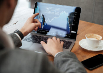 A person in a suit is engaged in day trading, using a laptop showing financial charts with varying trends. They're pointing at the screen, utilizing FintechZoom.com to make trading easier. A cup of coffee and a smartphone sit on the wooden table beside them. | FintechZoom