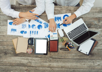 Two people at a wooden table with financial charts, a laptop, and tablets surrounded by various office supplies. One holds a pen while the other uses a calculator, showcasing their use of financial management tools. Notebooks, paperclips, and pens add to the busy work environment. | FintechZoom