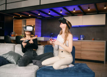 Two people sit on a couch in a modern living room, wearing virtual reality headsets and holding game controllers. One person wears black and sits comfortably, while the other is in white, sitting cross-legged on a blue cushion. | FintechZoom