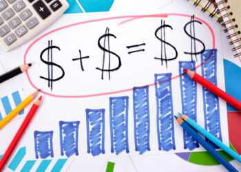 A paper with a hand-drawn equation: dollar sign plus dollar sign equals dollar sign, encircled in red. Below, a bar chart showcases rising blue bars—a vivid representation of financial literacy. Colored pencils, a calculator, and a notebook surround the paper. | FintechZoom