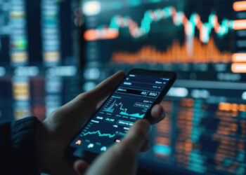 A person holds a smartphone displaying stock market data, surrounded by blurred computer screens with financial graphs. Glowing orange and blue elements indicate market trends while dApps seamlessly integrate into the digital display, showcasing innovative financial technology. | FintechZoom
