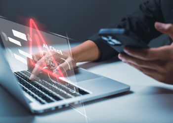 A person uses a smartphone to interact with a holographic AI chat interface projected from a laptop, seamlessly merging text bubbles and digital elements. This advanced technology symbolizes the future of connectivity, enhancing experiences like online banking against a blurred background. | FintechZoom