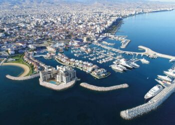 Aerial view of a coastline city with a marina filled with yachts and boats. The cityscape stretches into the distance, showcasing Limassol's property allure amid numerous buildings and a mountainous backdrop. The clear blue sea surrounds the harbor, featuring breakwaters and piers. | FintechZoom