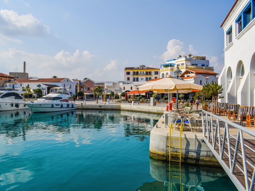 A marina with several docked boats on clear turquoise water. On the right, a restaurant with outdoor seating shaded by umbrellas. In the background, colorful buildings and a partly cloudy sky enhance the picturesque scene. | FintechZoom