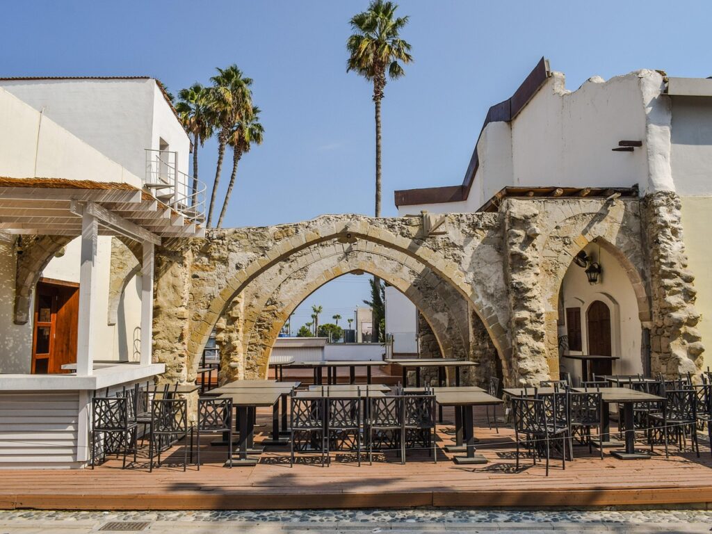 Outdoor seating area with empty black tables and chairs on a wooden deck. Surrounded by stone arches under a clear blue sky. Tall palm trees are visible in the background, enhancing the sunny atmosphere. | FintechZoom