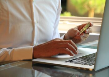 A person in a white shirt holding a credit card in their left hand while typing on a laptop with their right hand, seated at a table. Sunlight filters through a window in the background. | FintechZoom