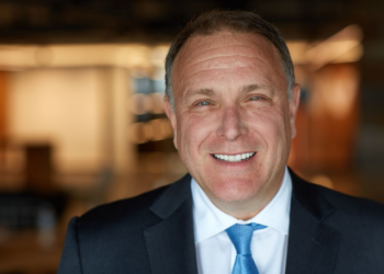Michael Polk in a suit and blue tie is smiling. The background is softly blurred, with warm lighting, giving a professional and friendly vibe. | FintechZoom
