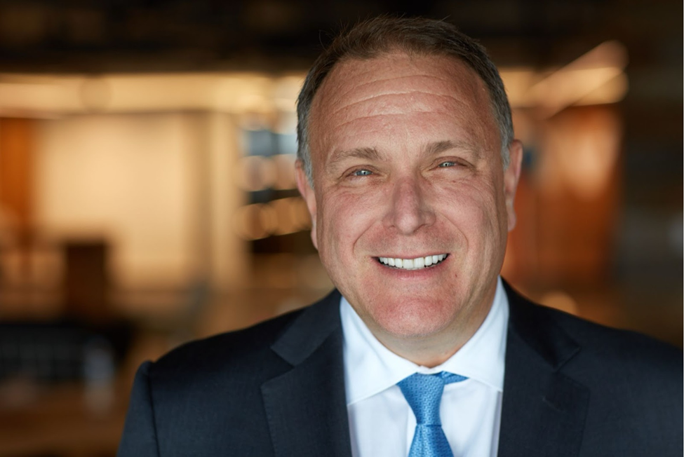 Michael Polk in a suit and blue tie is smiling. The background is softly blurred, with warm lighting, giving a professional and friendly vibe. | FintechZoom