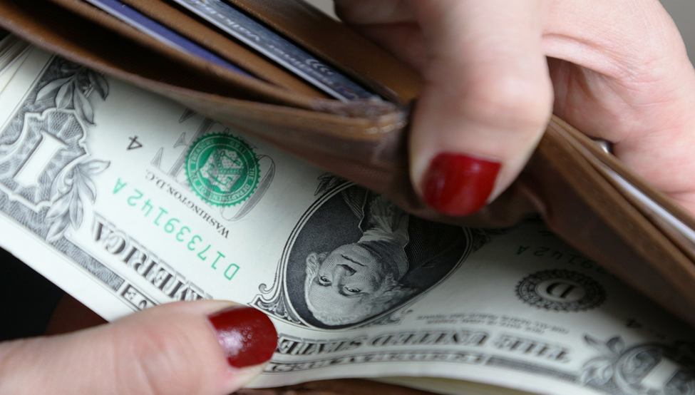 A person with red nail polish is holding an open wallet with a visible one-dollar bill. The focus is on the hands and the bill, highlighting the portrait of George Washington on the currency. | FintechZoom