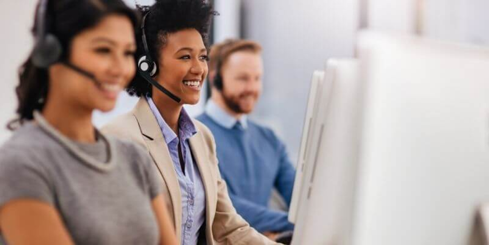 Three customer support representatives sit in a row, wearing headsets and smiling as they work at computer desks. The focus is on their positive expressions in a modern office setting. | FintechZoom