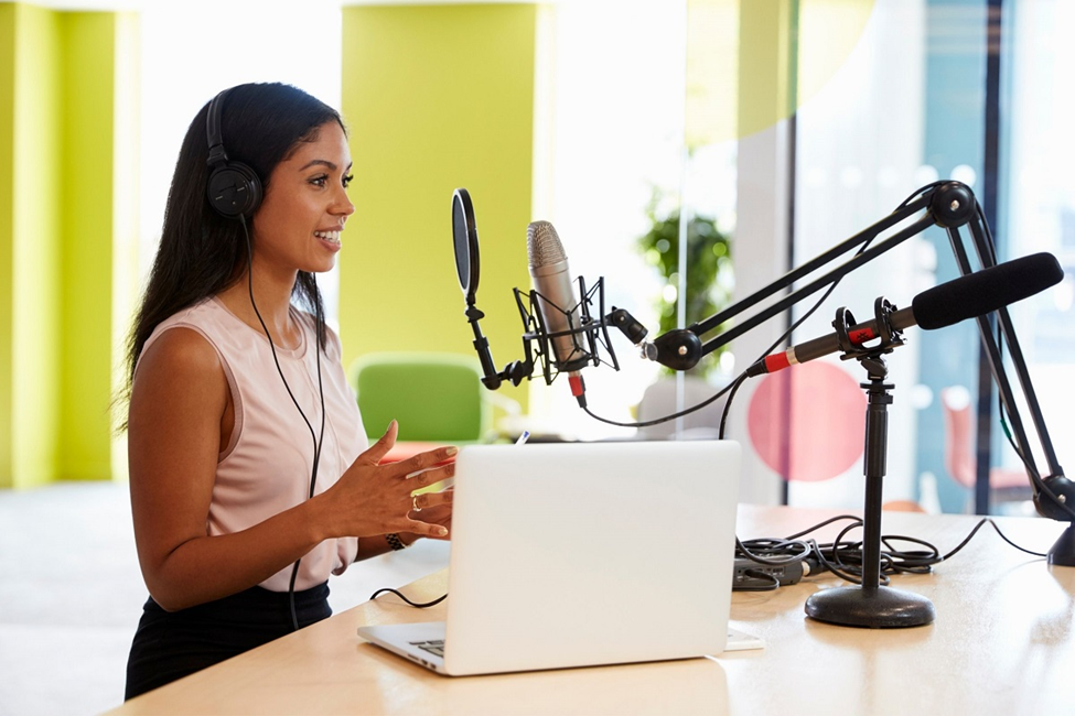 A woman wearing headphones speaks into a microphone at a desk with a laptop, utilizing AI Text-to-Speech for seamless narration. Surrounded by technical equipment like a boom arm and pop filter, she excels in her craft in a bright, modern studio environment. | FintechZoom