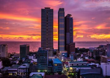A vibrant cityscape at sunset with two prominent skyscrapers silhouetted against a colorful sky of orange, pink, and purple hues. Below, various city buildings spread out towards the calm ocean on the horizon. | FintechZoom