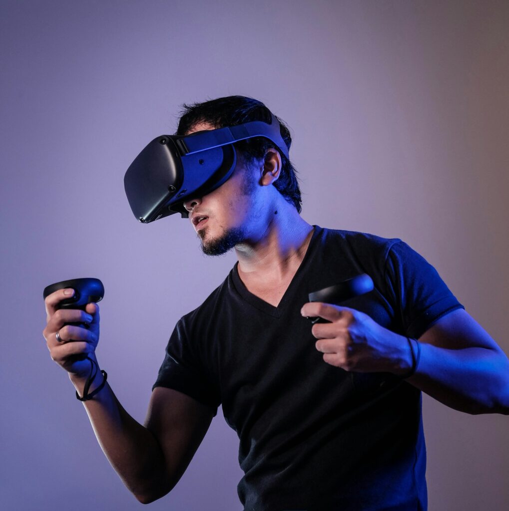 Person wearing a VR headset, holding controllers, appearing immersed in a virtual experience. He is dressed in a black t-shirt, set against a neutral background with blue lighting. | FintechZoom