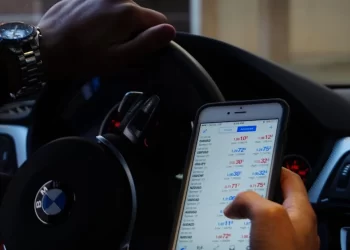 A person in a car holds a smartphone displaying a financial app with various currency exchange rates. Another hand is on the steering wheel. The car's interior is visible, including the steering wheel with a BMW logo. | FintechZoom