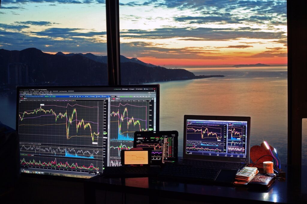 Multiple screens display stock market charts with line graphs and technical indicators. They are set up on a desk near a large window. A beautiful sunset view over the ocean is visible outside, with mountains silhouetted against the colorful sky. | FintechZoom