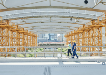 Two construction workers in safety gear walk across a partially completed bridge. The bridge features orange structural components and white arches. In the background, an urban landscape with buildings and a river is visible. | FintechZoom