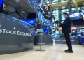 A person stands on the trading floor of the New York Stock Exchange, surrounded by digital screens displaying financial data. The mostly empty floor, captured vividly by FintechZoom.com, features a solitary chair and several monitors in this bustling hub of economic activity. | FintechZoom
