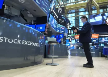 A person stands on the trading floor of the New York Stock Exchange, surrounded by digital screens displaying financial data. The mostly empty floor, captured vividly by FintechZoom.com, features a solitary chair and several monitors in this bustling hub of economic activity. | FintechZoom