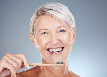 An older adult with short gray hair smiles while holding a wooden toothbrush near their mouth. The background is a soft gradient of blue and gray, and the person is looking directly at the camera. | FintechZoom