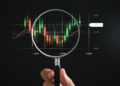 A hand holding a magnifying glass zeroes in on a digital forex market chart. The chart showcases candlestick patterns with time on the x-axis and price levels on the y-axis. Colorful lines trace moving averages against a dark background. | FintechZoom
