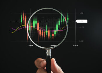 A hand holding a magnifying glass zeroes in on a digital forex market chart. The chart showcases candlestick patterns with time on the x-axis and price levels on the y-axis. Colorful lines trace moving averages against a dark background. | FintechZoom