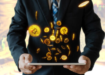 A person in a suit holds a tablet, from which glowing digital Bitcoin symbols appear to float upward, resembling the vibrant graphics of gaming. The blurred background with more Bitcoin imagery suggests a focus on cryptocurrency and technology that's being rapidly adopted. | FintechZoom