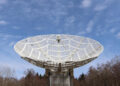 A large, white satellite dish points towards a clear blue sky, hinting at the ever-changing weather patterns. The dish is surrounded by leafless trees, indicating a winter or early spring setting. Wispy clouds are scattered across the sky. | FintechZoom