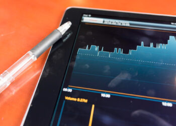 A tablet on a reddish-orange background showcases a line graph with fluctuating data and time indicators. Beside the tablet, a black pen with a transparent barrel rests. This scene is reminiscent of fintechzoom.com's insightful data presentations. | FintechZoom