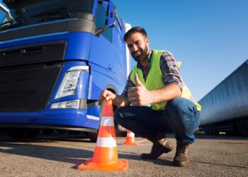 A man in a yellow safety vest crouches next to a blue truck, giving a thumbs-up, demonstrating safety awareness. He's holding an orange traffic cone under the clear blue sky, with a trailer in the background—ensuring measures against truck accidents on this sunny day. | FintechZoom