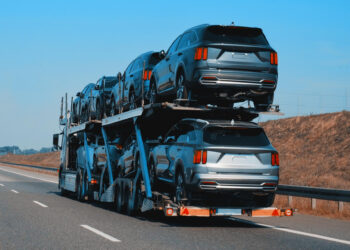A car carrier truck, crucial for financial market predictions for car shippers, transports multiple silver SUVs stacked on two levels along a highway. The road is clear under a bright blue sky, and dry grass lines the roadside. | FintechZoom