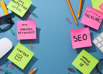 A blue surface adorned with colorful sticky notes illustrating SEO concepts such as "Backlinks," "Indexing Keywords," and "Site Architecture" highlights why SEO matters. Surrounding items like a keyboard, pencils, glasses, and paper clips complete the scene. | FintechZoom