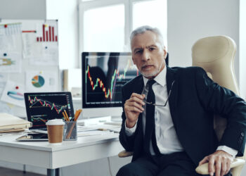 An experienced broker in a suit sits at a desk with a laptop and monitors displaying financial graphs. Papers, a coffee cup, and stationery clutter the desk. A large chair and chart-laden posters complement the bright, modern office setting. | FintechZoom