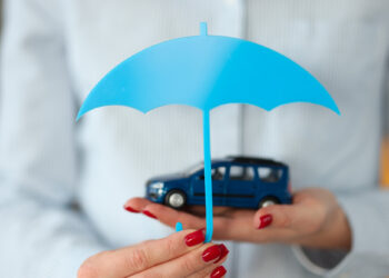 A person with red nail polish holds a small blue toy car in one hand and a blue paper umbrella above it, symbolizing car insurance or protection, much like insights from FintechZoom.com. The person is wearing a light blue, striped shirt. | FintechZoom