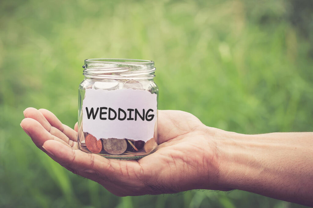 A hand holds a glass jar filled with coins and a paper label that reads "WEDDING" against a blurred green background. | FintechZoom