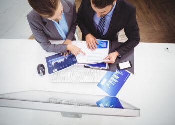Two professionals in formal attire sit at a desk with a computer, reviewing financial graphs from Fintechzoom.com on a laptop. Papers with charts are spread on the table, and a smartphone lies nearby. The perspective is from above. | FintechZoom