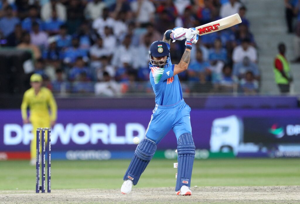 An India cricket player in a blue jersey and helmet elegantly plays a shot with his bat during the match. The enthusiastic crowd is visible in the blurred background. | FintechZoom