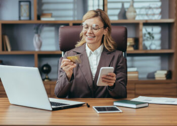 A woman in glasses and a business suit smiles while holding a credit card and smartphone, contemplating her next business loan online. Seated at a desk with a laptop, tablet, notebook, and papers, she is surrounded by the inspiration of bookshelves in the background. | FintechZoom