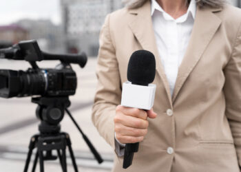 A person in a tan blazer is holding a microphone with a white square label, skillfully positioned to avoid disturbing wind noise. In the background, a video camera stands on a tripod, capturing the bustling outdoor urban scene. | FintechZoom
