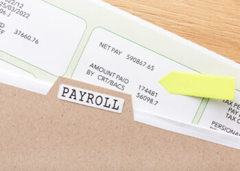 A close-up of a brown file folder labeled "Payroll" containing several payroll documents. One document shows details like pay period and amount paid. A small yellow arrow-shaped sticky note is attached to the file. The background is a wooden surface. | FintechZoom
