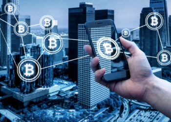 A hand holds a smartphone against a city skyline. The phone screen displays a connected network of Bitcoin symbols, represented by glowing icons, superimposed over skyscrapers, illustrating the world of crypto finance and digital transactions. | FintechZoom