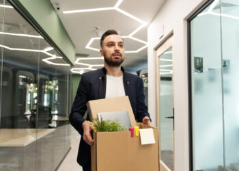 A man with a beard, donning a dark blazer, carries a cardboard box filled with a potted plant, a notebook, and other office items. As he strides down the modern hallway with glass walls and geometric ceiling lights, it’s clear he's in the process of relocating his business. | FintechZoom