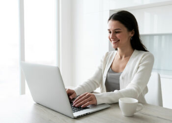 A woman sits at a table, smiling as she engages in a psychic reading on her laptop. Dressed in a white cardigan, with a white coffee cup beside her, she enjoys the serene atmosphere of the bright, minimalist room framed by neat shelves. | FintechZoom