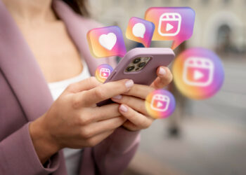 A person holds a smartphone with a lavender case, surrounded by colorful social media icons like hearts and speech bubbles, hinting at interactions or notifications. Clad in a mauve jacket, they're keen to purchase German Instagram followers to boost their online presence. | FintechZoom